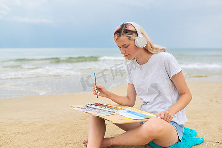 戴着耳机的年轻女子坐在沙滩上用水彩画素描
