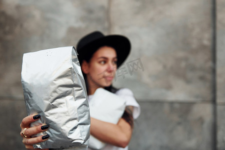 天气组件摄影照片_身穿黑帽和白衬衫的褐发女郎站在灰色墙壁附近，手里拿着带咖啡的包裹