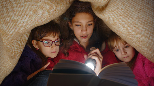 姐妹们在家里用手电筒在毯子下阅读有趣的童话故事书