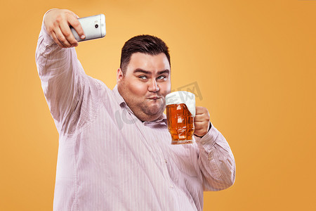 年轻的胖子在慕尼黑啤酒节上用黄色背景的啤酒在智能手机上制作自拍照片。