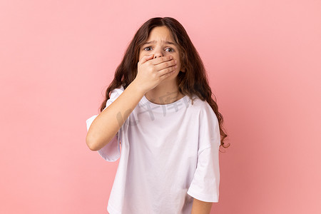 吓坏了摄影照片_穿着白色 T 恤的小女孩用手捂住嘴巴，保守秘密，被令人震惊的消息吓坏了。