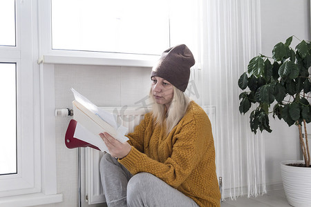 一个穿着黄色毛衣和一顶棕色帽子的年轻女孩坐在地板上，在带恒温器的加热器旁数钱，思考如何支付账单和税款。