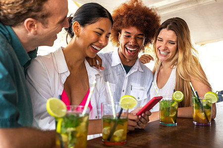 一群快乐的多种族朋友在海滩酒吧里看手机，喝着莫吉托鸡尾酒。
