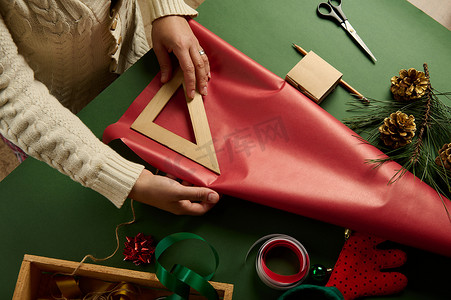 女人使用尺子，计算在绿色背景下包装圣诞礼物所需的红色包装纸的数量。