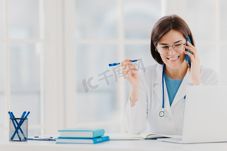高兴的专业医生专注于现代笔记本电脑，阅读有用的信息，进行电话交谈，讨论医疗问题，坐在医院办公室，桌上放着记事本