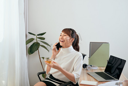 人喝茶摄影照片_年轻的亚洲女性一边喝茶一边坐在办公室里打电话