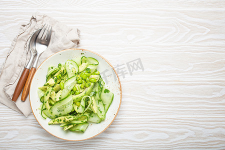 健康的素食绿色鳄梨沙拉碗，配有切片黄瓜、毛豆、橄榄油和香草，陶瓷盘顶视图，白色木制乡村餐桌背景。