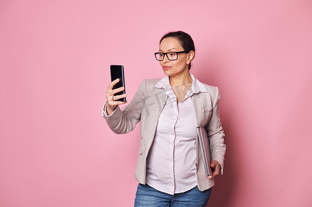 怀孕自信的西班牙裔女商人拿着笔记本电脑，在她的智能手机上查看移动应用程序，孤立的粉红色背景