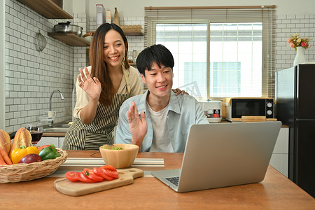 舒适家人摄影照片_微笑的亚洲男人和妻子打视频电话，在现代厨房的笔记本电脑上与家人或朋友在线聊天