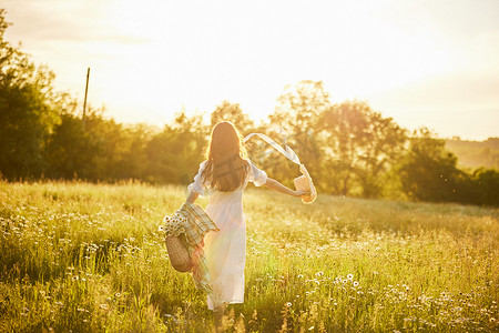 一位穿着浅色连衣裙的女士站在田野里，沐浴在夕阳的温暖光线中，头上戴着一顶帽子