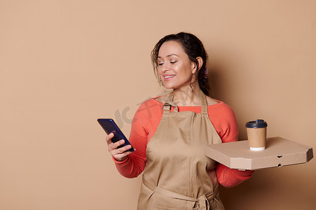 纸杯样机摄影照片_身穿厨师围裙、微笑的女服务员拿着手机、带热披萨的纸板盒和纸杯中的外卖咖啡