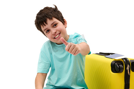带着黄色手提箱的快乐的青春期前乘客男孩，竖起大拇指，对着镜头微笑，孤立的白色背景