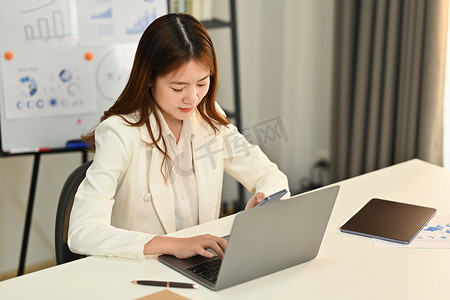 专注的亚洲女经济学家看着笔记本电脑屏幕检查电子邮件，在她的办公桌上处理统计数据