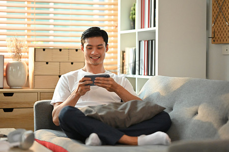坐在家里的沙发上，随意观看视频或在智能手机上玩游戏的快乐亚洲男人