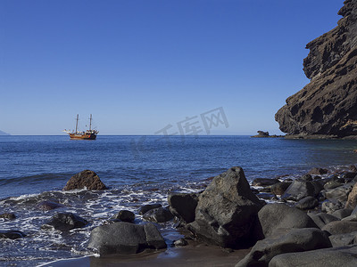 从黑沙滩 Playa De Masca 与大石头、悬崖和蓝天背景的旧帆船欣赏海平面