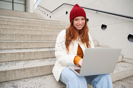 微笑的红发女孩，在笔记本电脑键盘上打字的年轻女性，坐在户外楼梯上用电脑，远程工作，在新鲜空气中做作业