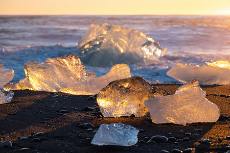 纯冰在日落时在黑色火山沙滩上闪耀，海浪冲撞冰山