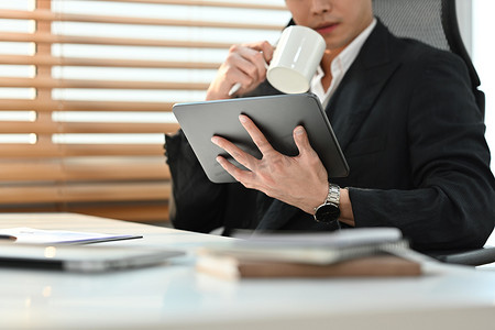 身穿黑色西装的男子坐在办公室窗边喝咖啡并在数字平板电脑上阅读在线新闻的剪影