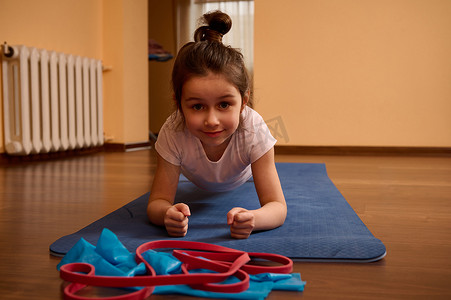 坚定的运动小女孩在蓝色健身垫上做平板运动，同时在室内锻炼和伸展身体
