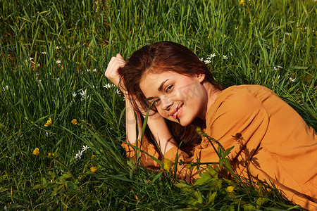 一个长着红色长发的平静女人躺在开着黄色花朵的绿色田野里，身穿橙色连衣裙，愉快地微笑着，在灿烂的阳光下眯着眼睛看着镜头