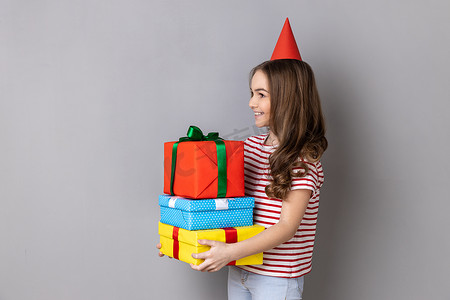 快乐迷人的小女孩穿着条纹 T 恤和派对锥，拥抱着一堆礼物。