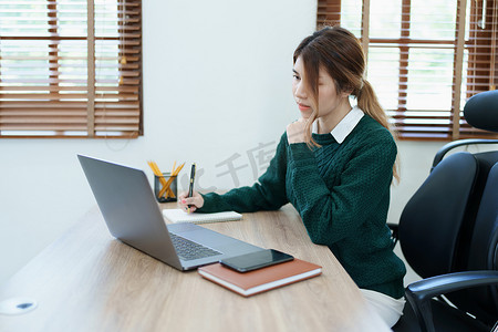 一位年轻亚洲女性的画像，她在清晨使用办公桌上的笔记本、电脑和财务文件时面带微笑