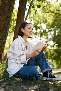 在温暖的阳光明媚的日子里，快乐的亚洲女性在公园户外放松、阅读她的书并坐在树荫下的垂直照片