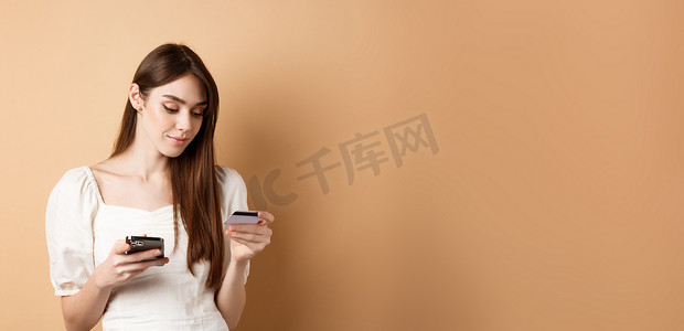 购物app摄影照片_微笑的年轻女性在线支付，查看信用卡并在手机上购物，在互联网上购物，站在米色背景中