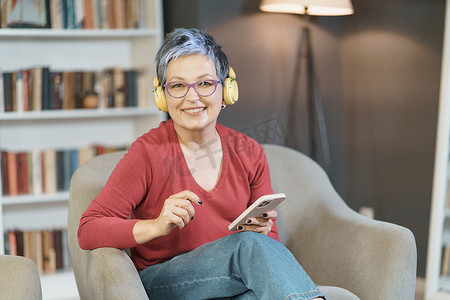 高级女士在家里戴着粉色眼镜和黄色耳机，正在愉快地用手机发短信，反映出一种注重技术和交流的轻松现代生活方式。