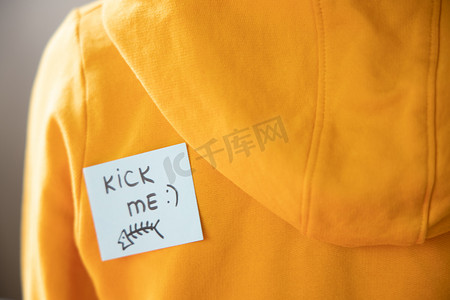 愚人节活动背景摄影照片_身穿黄色连帽衫的男孩背上贴着便利贴，上面写着踢我。