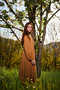 一位长着红色长发的甜美迷人的女人穿着橙色长裙站在一棵开花的树旁，脸上带着淡淡的微笑看着镜头