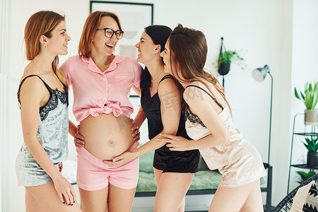 快乐的年轻女性和穿着睡衣的怀孕朋友站在一起，白天在室内聚会