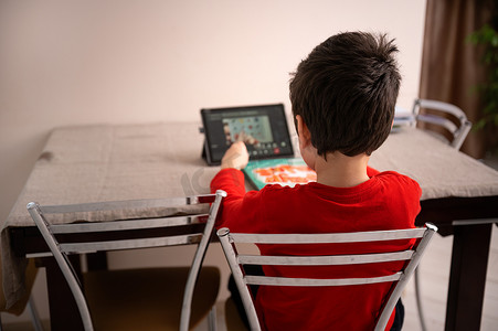 一个学童做作业、坐在桌边、在数字平板电脑上观看在线课程的后视图