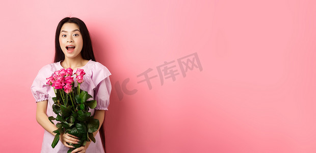 韩国少女穿着裙子在情人节浪漫约会，手捧玫瑰花束，对着镜头看起来很惊讶，在约会时收到情人送的礼物，粉红色背景