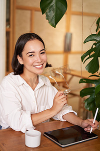 美丽的亚洲女孩坐在咖啡馆里喝杯咖啡，用图形笔在数字平板电脑上画画，轻而易举地画画