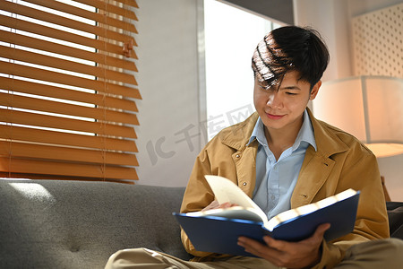 轻松的千禧一代男人在舒适的沙发上看书，在家享受休闲周末
