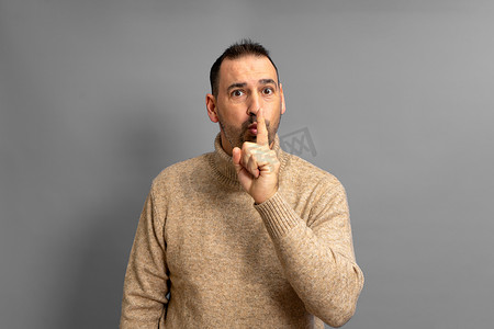 西班牙裔英俊男子身穿米色高领毛衣，身穿孤立的灰色背景，手指放在嘴唇上要求保持沉默。