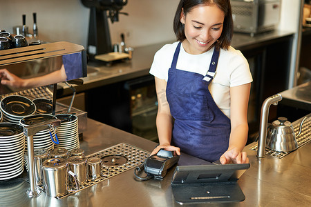 在咖啡厅柜台工作、处理订单、使用 POS 终端接收咖啡付款的微笑亚洲女咖啡师