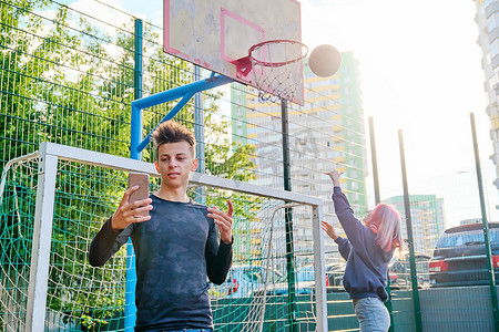 男孩青少年使用智能手机录制视频流，在体育街球场上，女孩打篮球