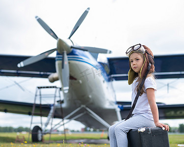 一个穿着飞行员服装的小女孩坐在机场的复古手提箱上等待航班起飞。