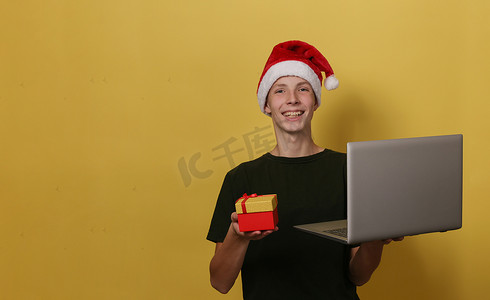 戴着圣诞帽的迷人 15 岁男孩拿着灰色笔记本电脑和红色礼盒。