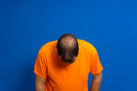 身穿橙色 T 恤的男人向前倾着头，露出他的秃头，与蓝色工作室背景隔离。