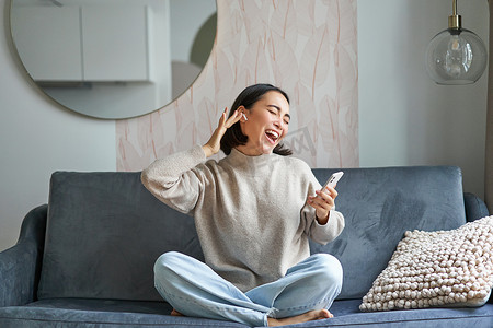 坐着听歌摄影照片_无忧无虑的亚洲女性在智能手机应用程序中唱歌和听音乐的肖像，使用无线耳机，微笑着高兴，坐在家里的沙发上