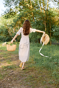 飘逸长裙摄影照片_一位身穿夏日长裙的女士在林间小径上行走，手里拿着一顶饰有飘逸缎带的柳条帽