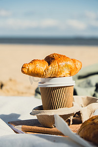 美味的新鲜羊角面包和沙滩海岸背景纸杯中的咖啡。