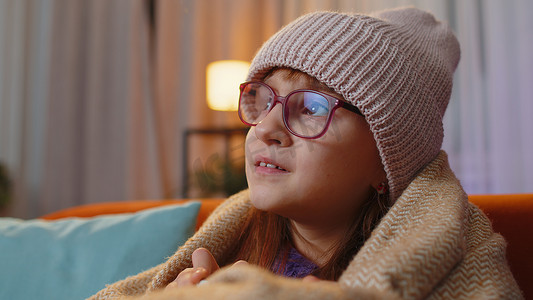 花呢摄影照片_生病的蹒跚学步的女孩戴着裹着格子花呢的帽子，独自坐在沙发上喝着热可可因寒冷而发抖