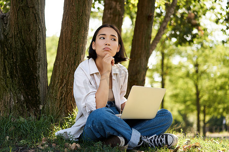 坐在树边公园里、在笔记本电脑上工作、在户外使用电脑的亚洲年轻女性的画像
