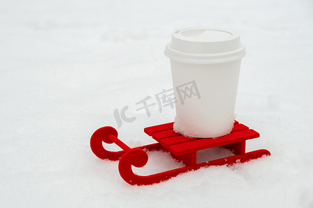 在美丽的冬日，雪背景上带纸杯咖啡或热巧克力的小红色雪橇。