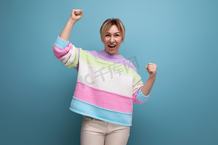 条纹毛衣摄影照片_身穿条纹毛衣的可爱迷人金发年轻女性在蓝色背景中表现出快乐的幸福情绪