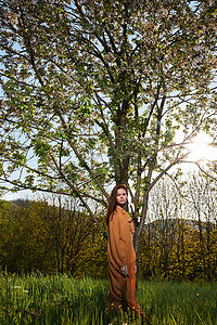 绿色树苗条摄影照片_一位身材苗条、冷静、留着长长的红头发的女人站在一棵开花的树旁，身穿橙色长裙，她看着镜头，享受着寂静